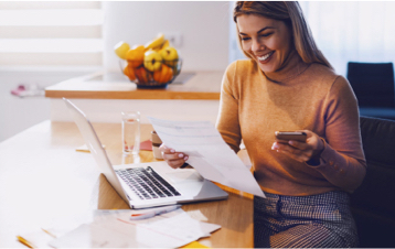 Una donna seduta a un tavolo con un portatile, che tiene in mano un telefono e un foglio.